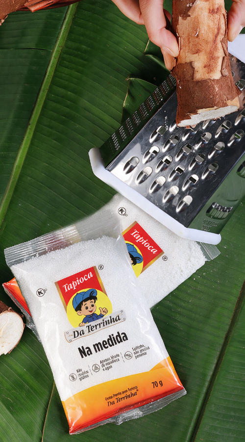 Mandioca sendo ralada sobre folhas de bananeira, ao lado de embalagens de goma pronta para tapioca Da Terrinha